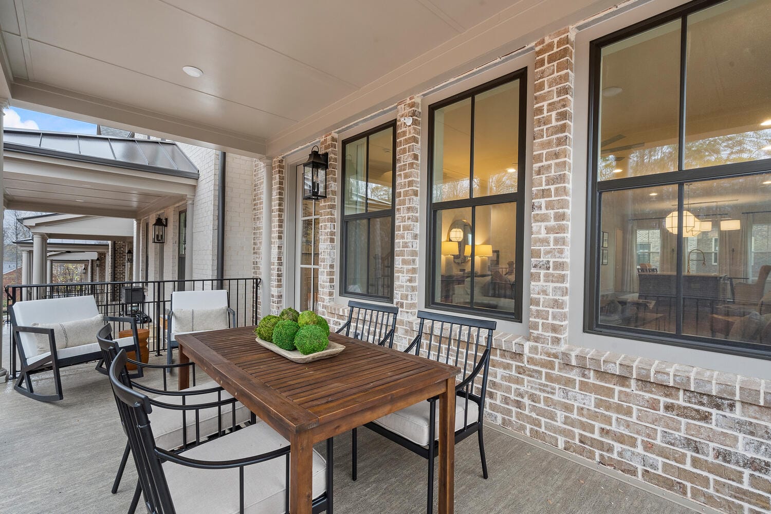 Outdoor patio area attached to a house with brick walls, wooden table with black metal chairs, rectangular tray holding green decorative spheres, covered roof, large windows providing view into interior with lights and furniture, white cushioned seating areas along patio railing.