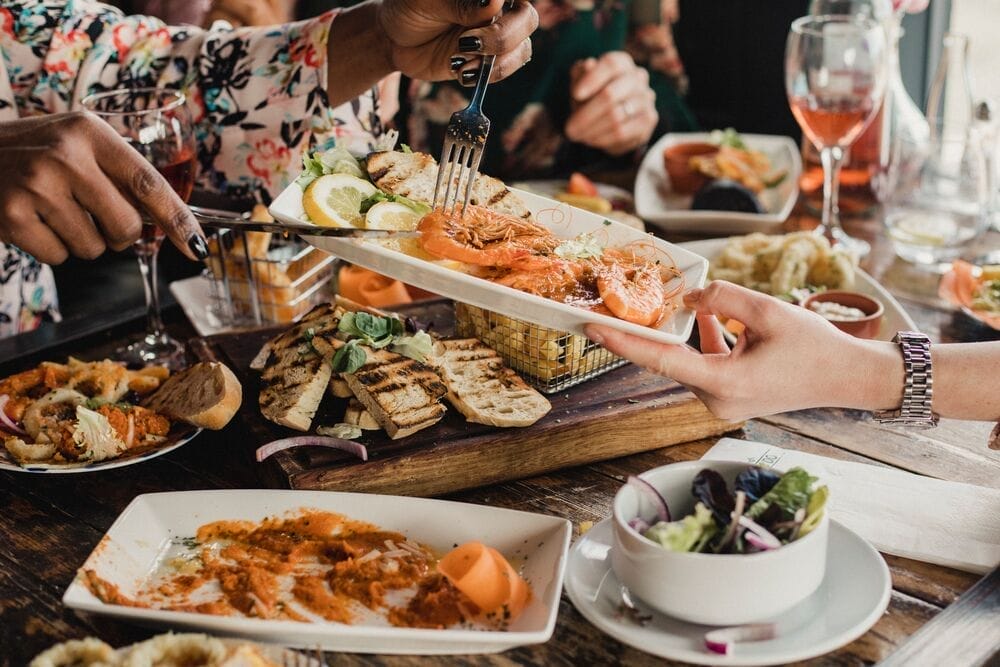 Point of view image of a group of multi-ethnic friends sharing multiple dishes, with a plate of prawns as the main focus.