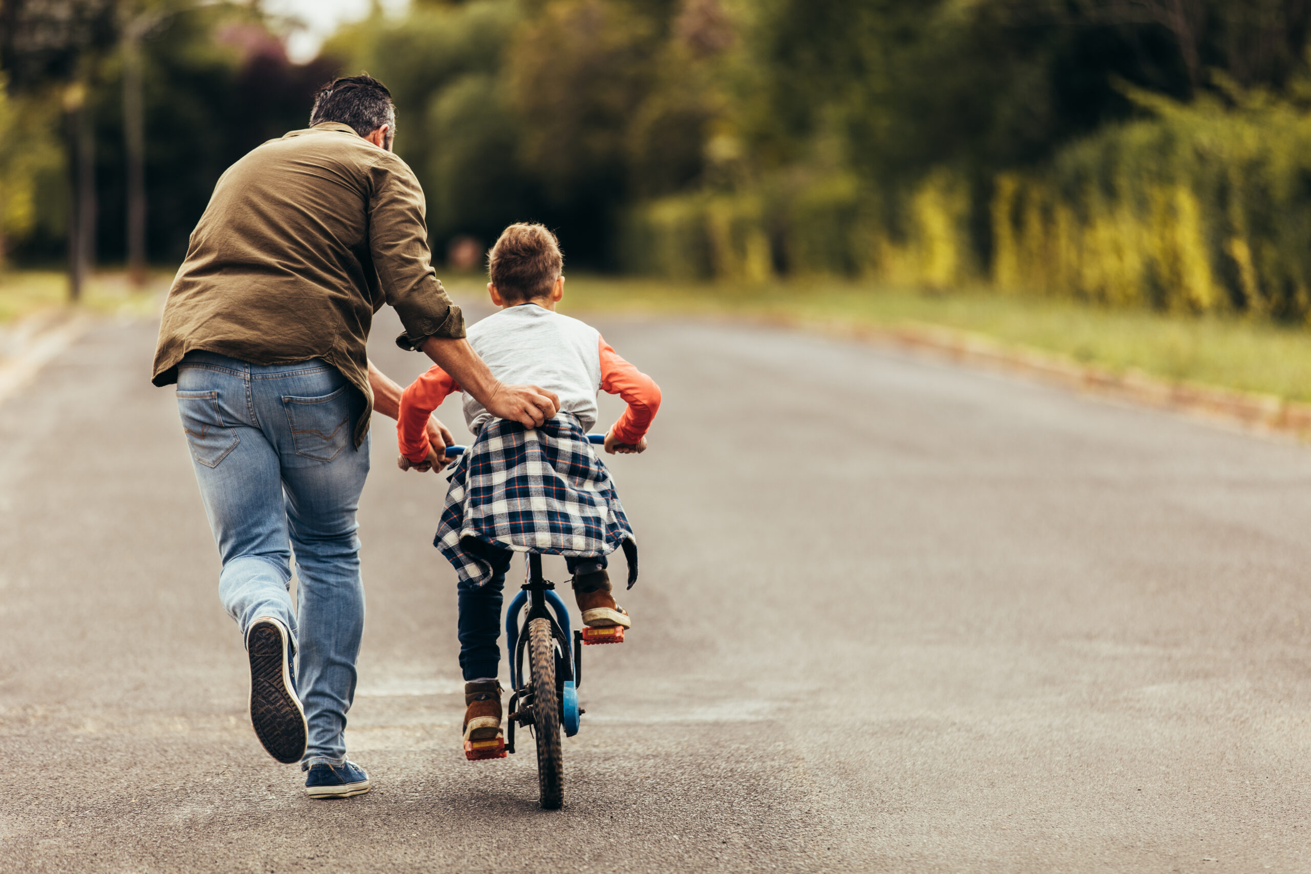 Rear view of a boy riding a bicycle while his father runs along holding the kid. Father teaching his son to ride a bicycle.