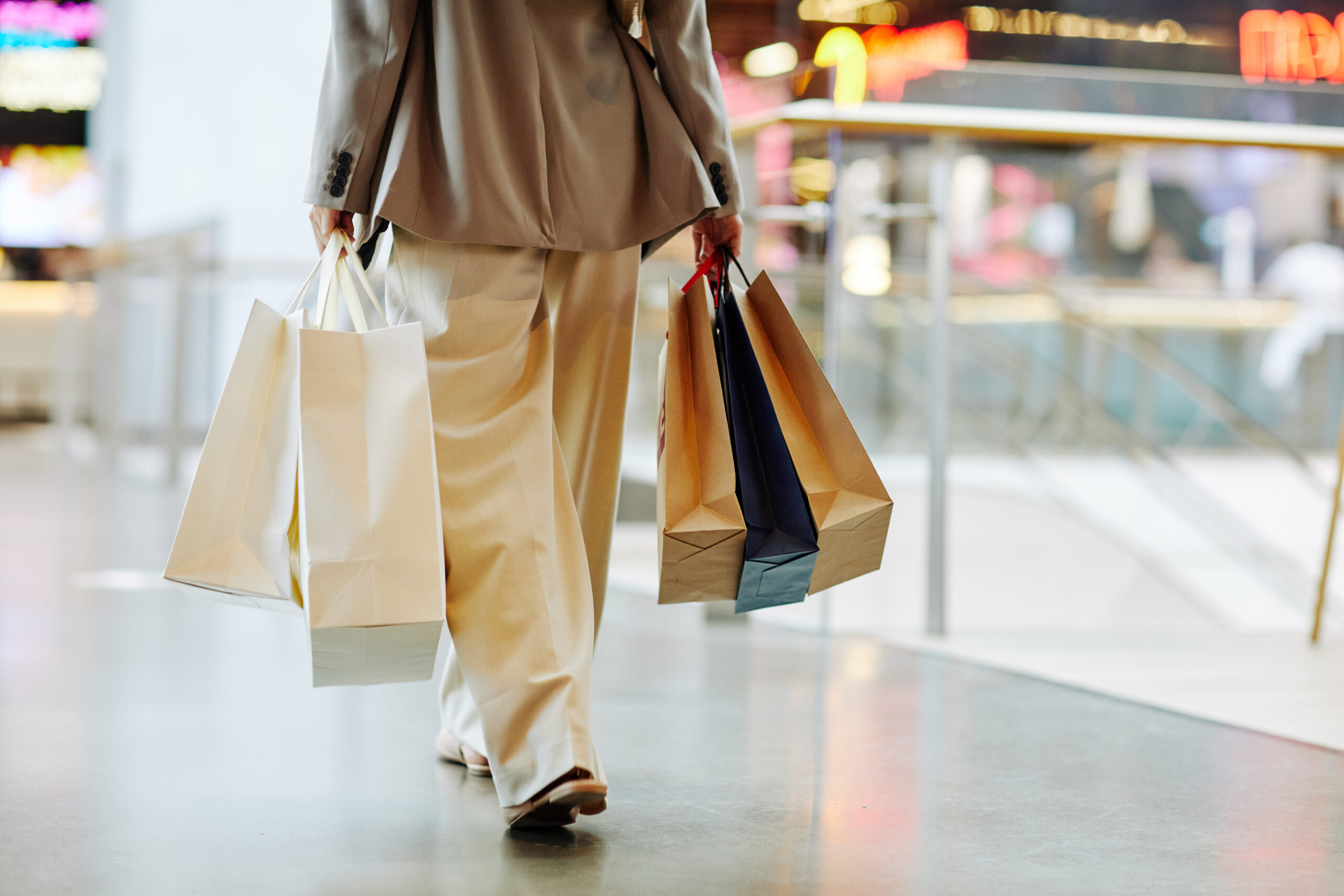 Low section of unrecognizable woman wearing pants and holding blank shopping bags while walking in mall, copy space
