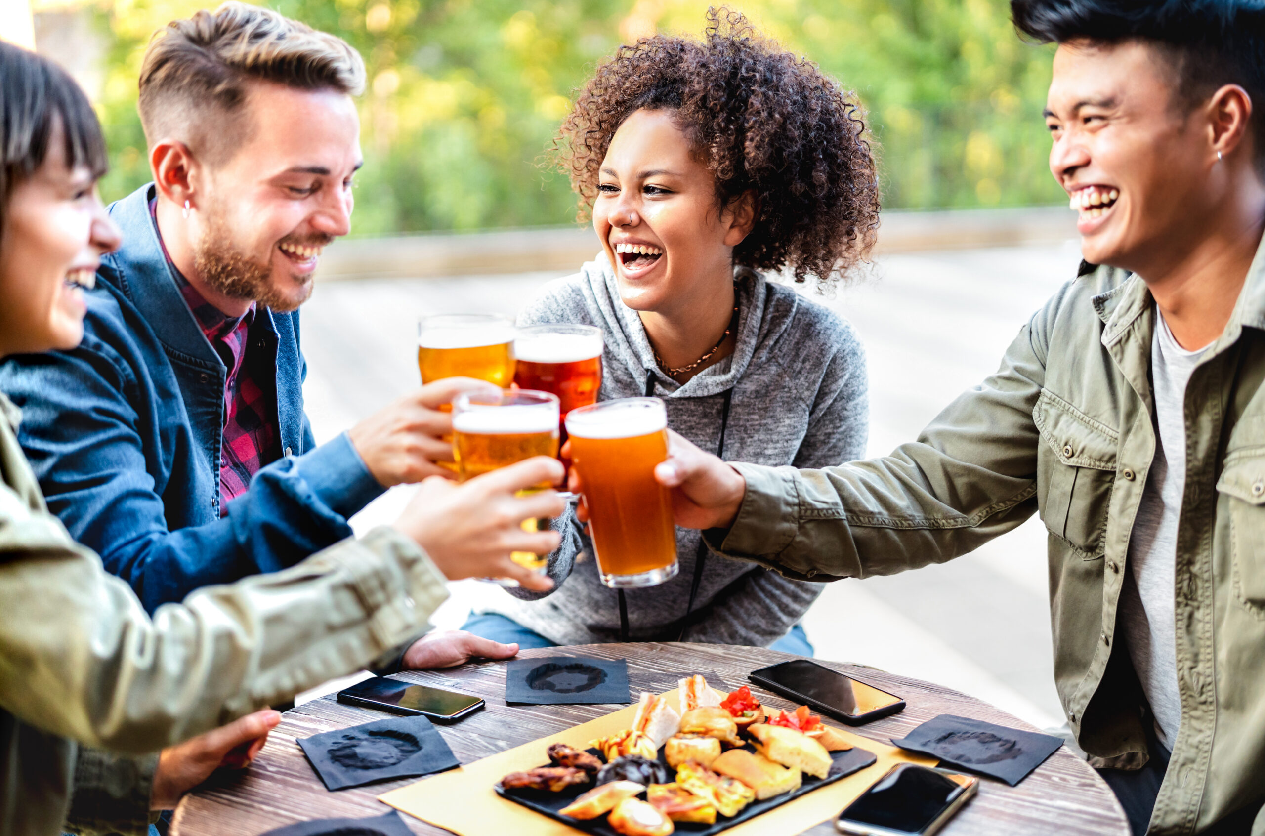 Young multicultural friends drinking and toasting beer at brewery bar patio - Youth life style concept with men and women having fun together out side - Bright vivid filter with focus on mid girl