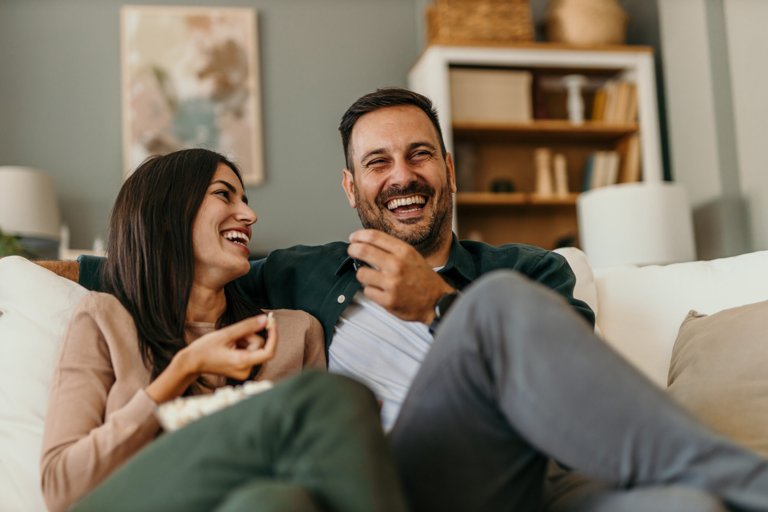 Happy young couple watching tv in the living room of their home and laughing hard